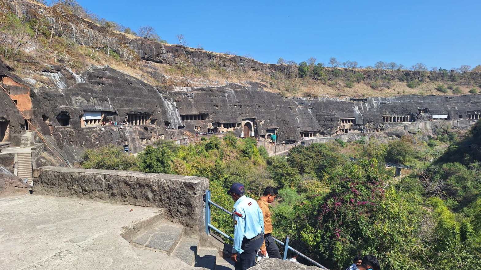 Panoramic view of the Ajanta Caves carved into a curved basalt cliff face in Maharashtra, India, with visitors on foreground stone steps descending toward lush green vegetation and the cave complex beyond.