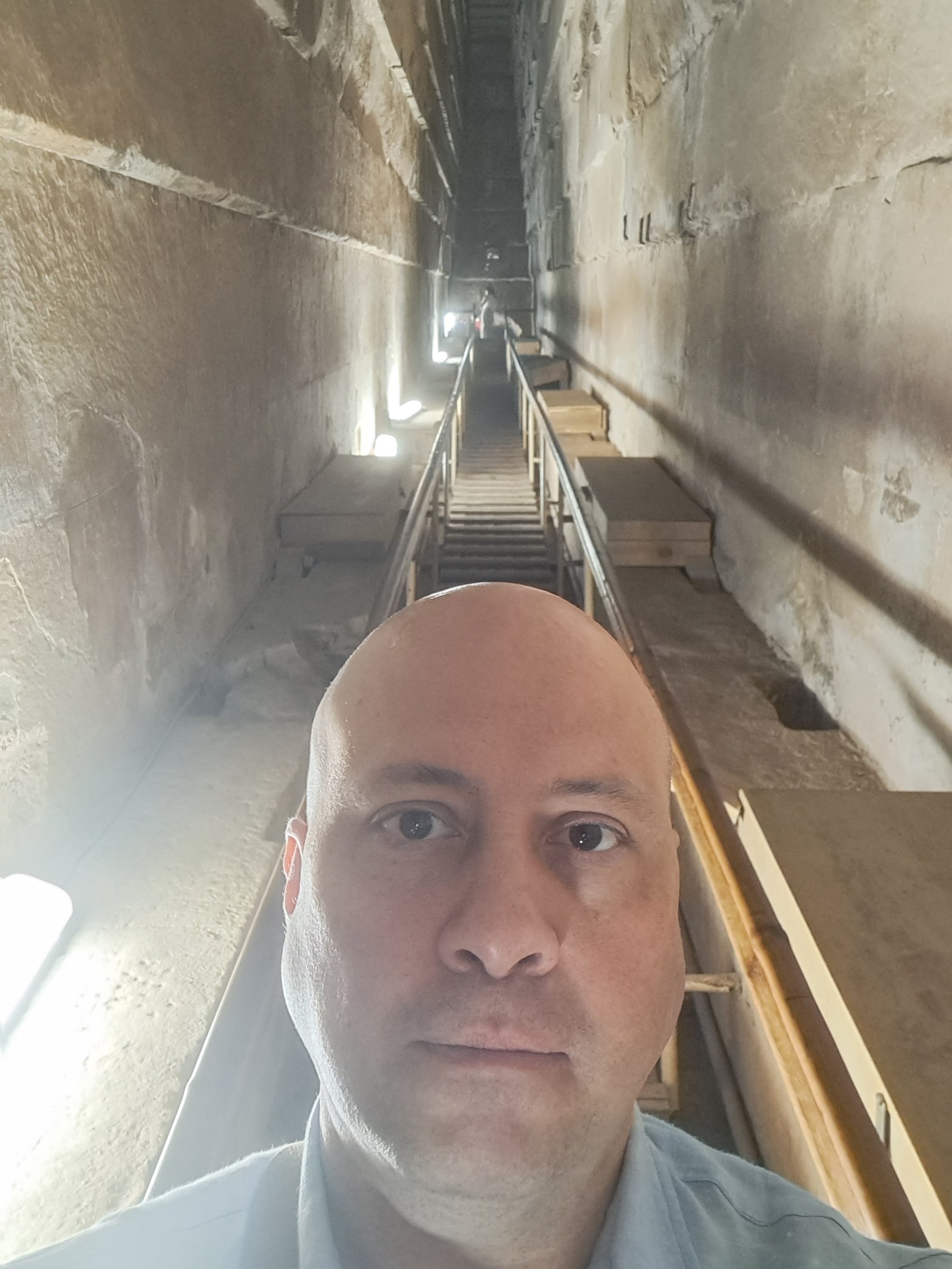 A man takes a selfie inside a narrow ascending passage within the Great Pyramid of Giza. Ancient limestone walls line both sides. A wooden staircase with metal railings recedes toward a lit opening in the distance.