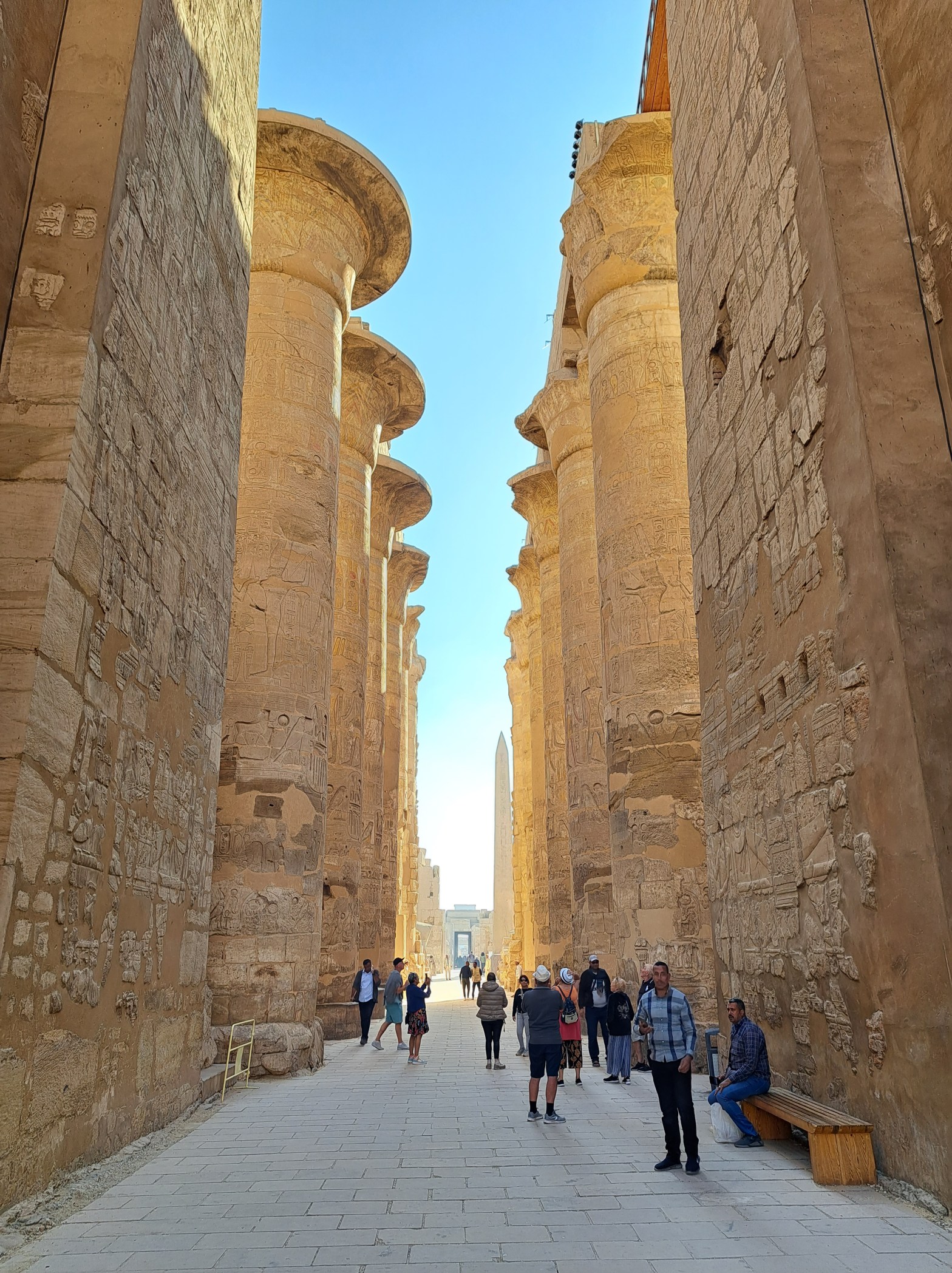 Massive stone columns of the Great Hypostyle Hall at Karnak Temple in Luxor, Egypt, covered in hieroglyphic carvings, with tourists walking beneath them and an obelisk visible in the distance.