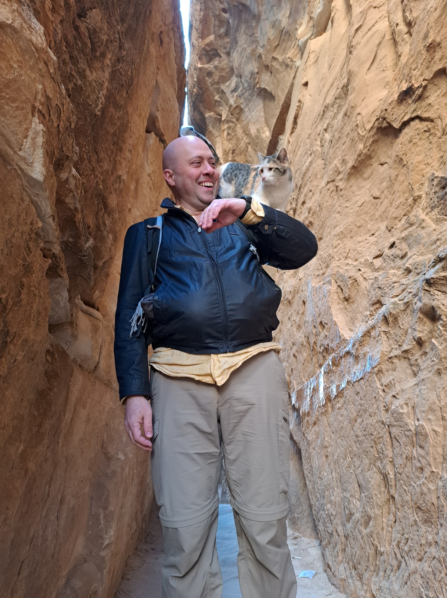 A bald man in a black jacket smiles while standing in a narrow sandstone canyon at Petra, Jordan. A gray and white cat perches on his shoulder behind him, alert and calm.