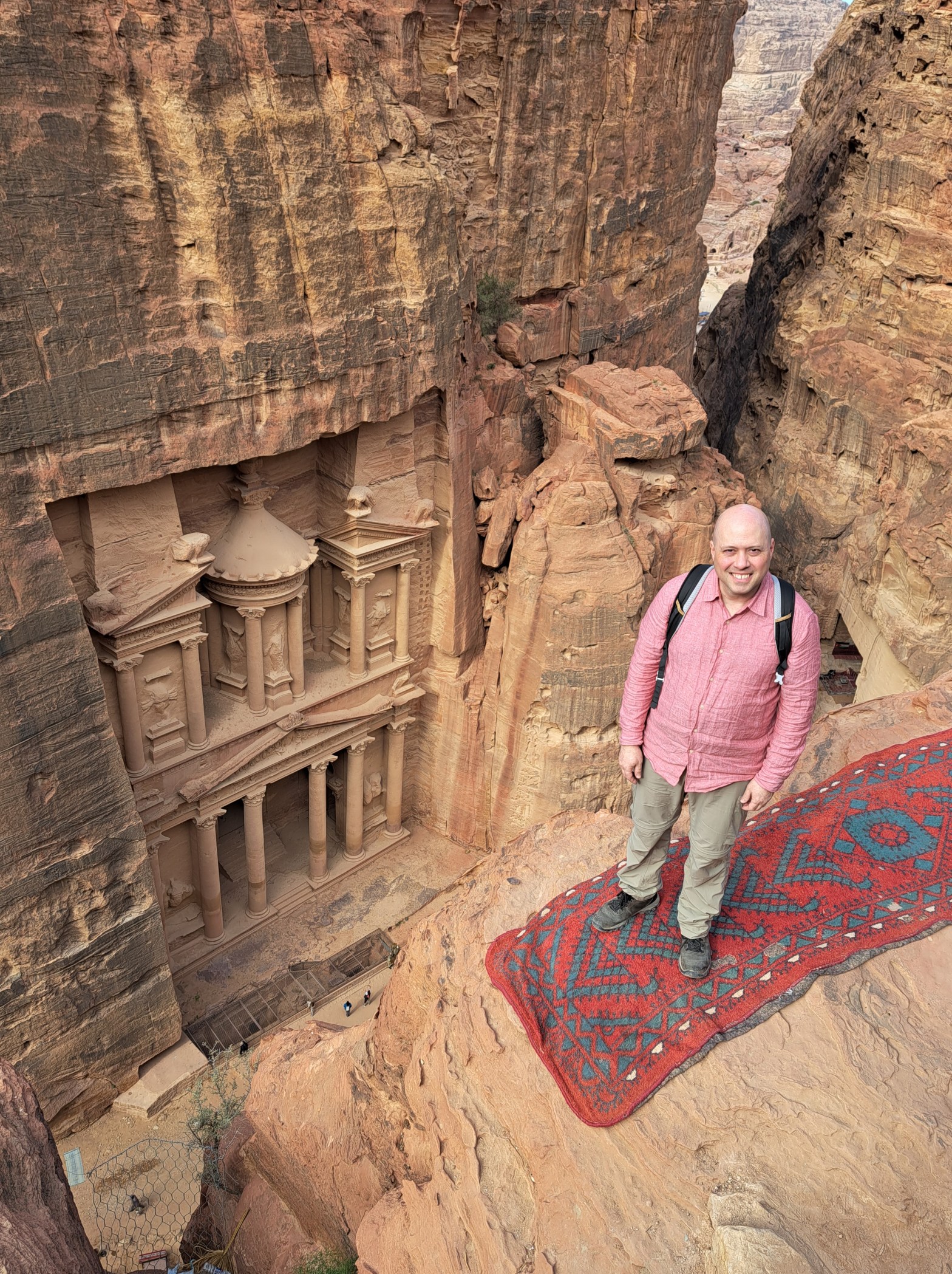 Man standing on a sandstone ledge above Petra's Treasury facade, carved into rose-red cliffs in southern Jordan, with a traditional red and teal Bedouin rug beneath his feet.
