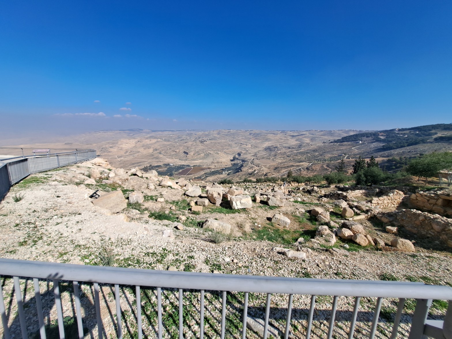 View from the summit of Mount Nebo in Jordan, looking westward across a vast arid valley and rolling hills toward the land of Canaan, with ancient stone ruins in the foreground and a clear blue sky above.