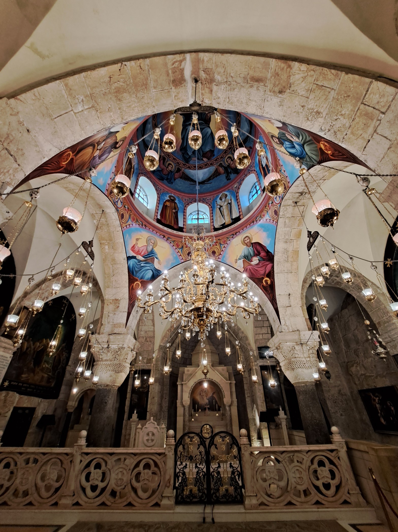 Interior of the Church of the Holy Sepulchre, Jerusalem, looking upward through a stone archway toward a painted dome depicting Christ and saints, with ornate gold chandeliers hanging in the foreground and an iron gate sanctuary screen below.